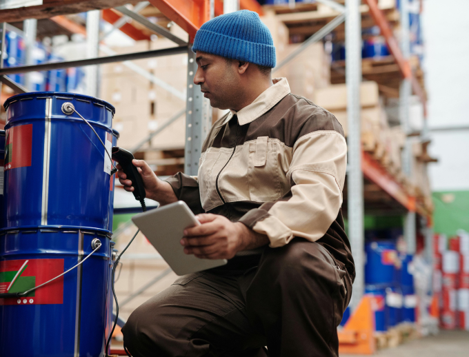 A man in aa blue hat with brown and tan work clothes kneels down in a warehouse to scan a large blue metal bucket.